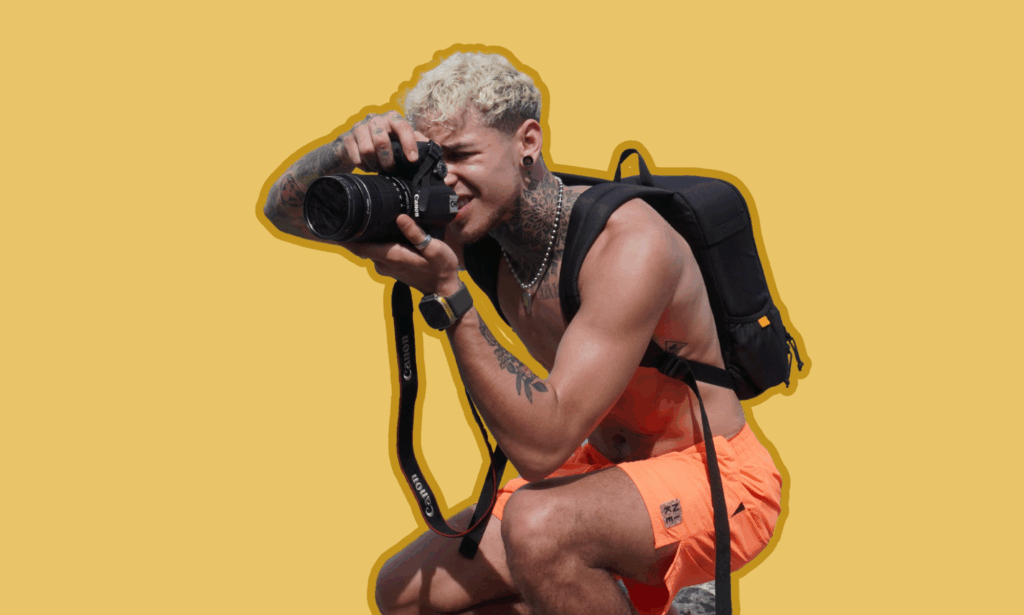Dominic Pérignon, shirtless and wearing bright orange shorts, crouches with a Canon camera in hand, aiming through the viewfinder with focus. He wears a black backpack and jewelry, framed against a golden yellow background with a soft brown outline.