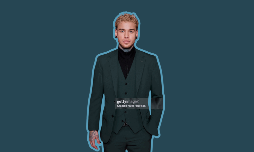 Dominic Pérignon poses on the red carpet at the GLAAD Media Awards in a dark green suit layered over a black zip-up shirt, outlined in light blue against a deep teal background, with visible tattoos and platinum curls.