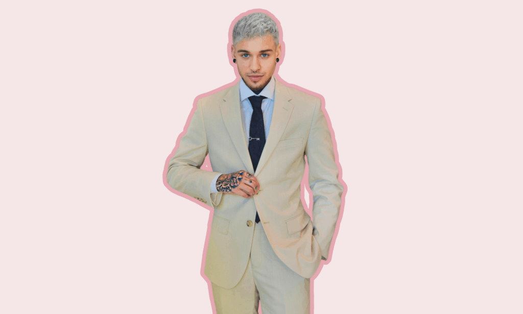 Dominic Pérignon poses confidently in a tailored beige suit from SuitShop, standing against a soft pink background with a pink outline effect, showcasing his hand tattoos and platinum hair.